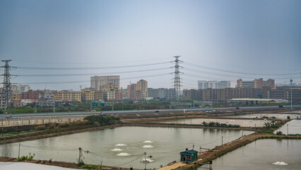 Fototapeta premium Between Guangzhou, Canton, and Macao, China, 3 March 2025: view from train showing aquaculture ponds, highway and residential blocks. Rapid urban growth in the Pearl River Delta.