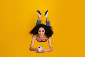 Young woman with curly hair wearing casual chic style smiles while holding a smartphone against a...