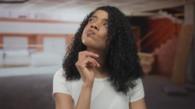 Woman with curly hair resting hand on chin inside unfinished building with exposed brick walls and scattered lumber; contemplation.