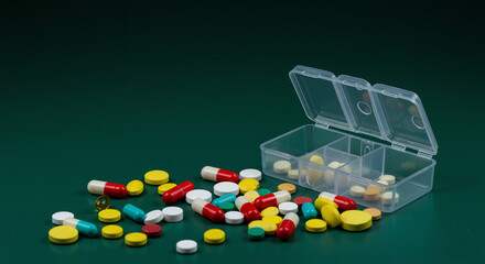AssoA scattering of colorful vitamins and capsules on a dark green background next to an open plastic medicine organizer. Each capsule is a vivid smear in the daily picture of your health, requiring p