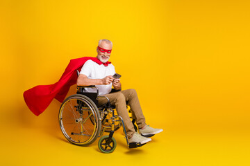 A cheerful grandpa in wheelchair wears a red cape and mask while using a phone against a bright yellow backdrop
