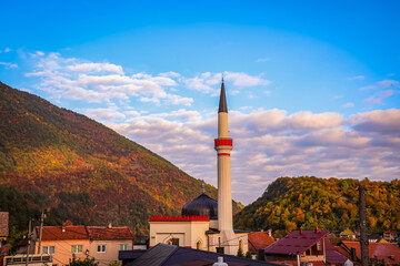 Fototapeta premium Beautiful mosque with tall minaret surrounded by autumn-colored hills and village houses in Travnik, Bosnia and Herzegovina, under a bright blue sky with scattered clouds.