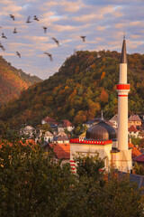 Beautiful autumn view of Travnik, Bosnia and Herzegovina, with a mosque minaret and dome among colorful hills and traditional houses, birds flying under a soft evening sky.