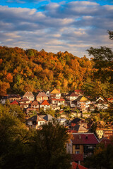 Naklejka premium Picturesque autumn view of Travnik, Bosnia and Herzegovina, with hillside houses glowing in warm evening light surrounded by colorful forest under a partly cloudy sky.