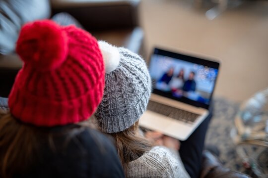 Two individuals wear knitted winter hats, watching Christmas video call on laptop screen in cozy room.
