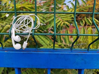 Tangled White Wired Earbuds with Lightning Connector, Hanging on a Green Metal Mesh Fence Above a Blue Rail, Against a Backdrop of Green Foliage.