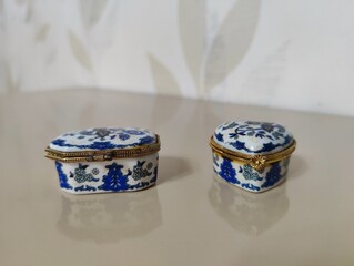 Pair of Small Vintage Porcelain Trinket Boxes with Blue and White Floral Patterns and Brass Clasps, Resting on a Reflective Beige Tabletop with Soft Background.