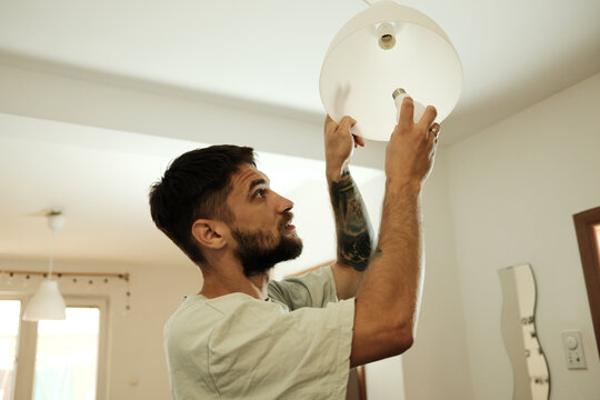 Young man with beard and tattoos installing a new light bulb in a ceiling lamp inside a bright modern apartment. Concept of home maintenance and everyday domestic life