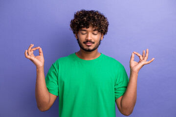 Relaxed young man in a green t-shirt meditating with a calm expression and hand gesture against a violet background