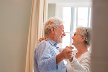 Joyful active old retired romantic couple dancing laughing in living room, happy middle aged wife and elder husband having fun at home, smiling senior family grandparents relaxing bonding together