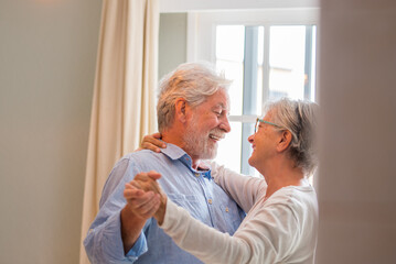 Joyful active old retired romantic couple dancing laughing in living room, happy middle aged wife and elder husband having fun at home, smiling senior family grandparents relaxing bonding together