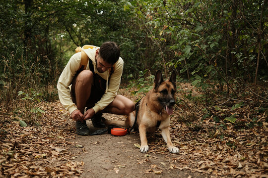 Young man in hoodie tying his shoelace on a forest trail while his German Shepherd dog waits beside him. Concept of outdoor recreation and hiking with pets - Powered by Adobe