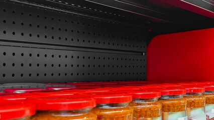 Rows of crimson-lidded jars linger in suspense on an empty shelf, evoking pantry preparation for...