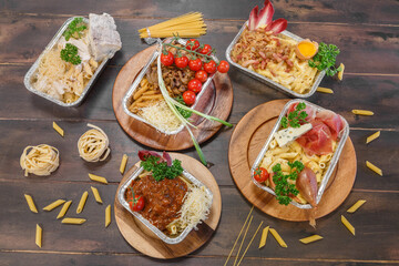 Pâtes à emporter – assortiment de pâtes bolognaise, carbonara, 4 fromages, jambon, roquefort, stylisées sur une table en bois avec décorations, vue 90°. Plats à emporter. 