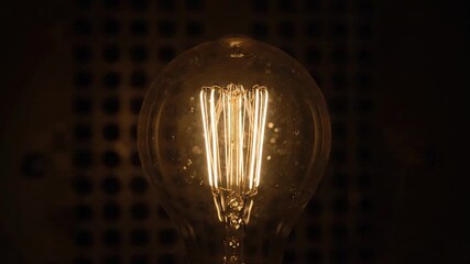 Glowing lightbulb, filament illuminated inside a clear glass with a dark patterned background - Powered by Adobe