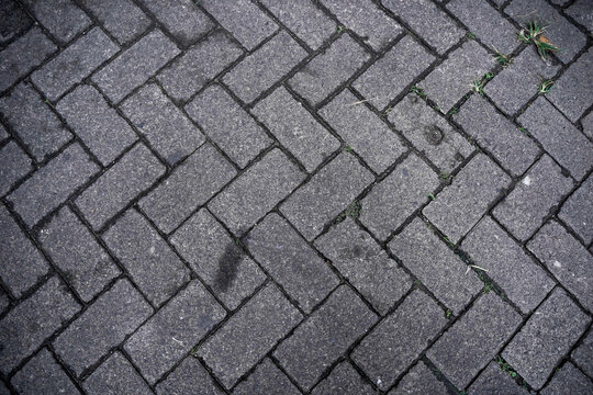 Top-down view of grey paving stones in a herringbone pattern. Pavement block texture background with weeds