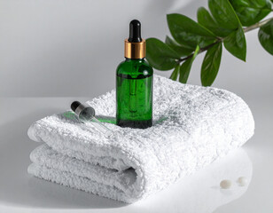 Green glass cosmetic dropper bottle for serum or essential oil on a folded white towel with a houseplant.