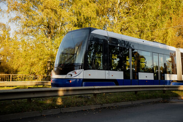 Fototapeta premium Modern blue and white electric tram moving along city track surrounded by autumn trees in Riga, showing contemporary public transportation design and sustainable urban mobility