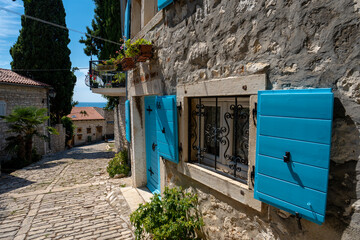 Rovinj, Croatia - August 14, 2025: Charming cobblestone street lined with stone houses featuring bright turquoise shutters, iron-grilled windows, and small balconies with flowers under clear sunny ski
