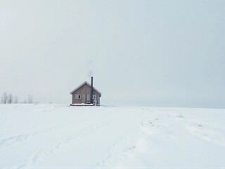 A small wooden cabin stands alone in a vast, snow-covered field under a cloudy, overcast sky, with smoke rising from its chimney.
