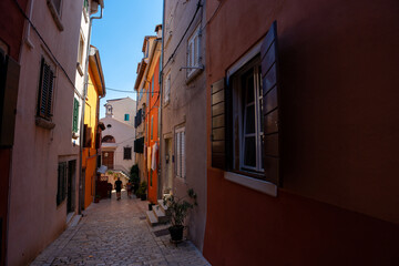 Rovinj, Croatia - August 14, 2025: A narrow cobblestone alley lined with pastel walls leads toward a small chapel with a bell tower, glowing under the midday sun.