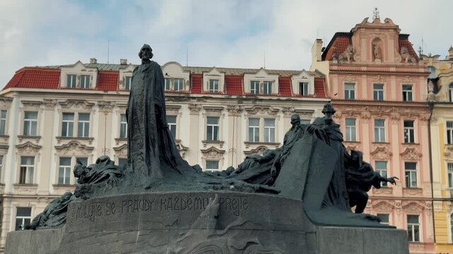 The historic Jan Hus Monument stands in Prague's Old Town Square, surrounded by baroque architecture. Tourists and locals walk near the iconic bronze statue.

