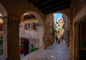 Fototapeta premium Rovinj, Croatia - August 14, 2025: Arched passageway opens to a sunlit cobblestone street lined with rustic walls and wooden shutters, where a small family strolls through the quiet charm