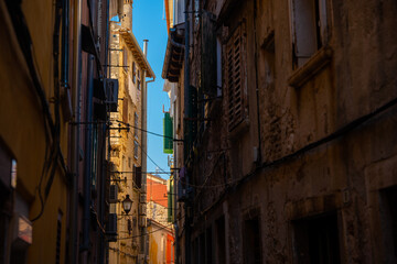 Fototapeta premium Rovinj, Croatia - August 14, 2025: Colorful wooden shutters and drying laundry line a narrow old-town alley, where sunlight paints the weathered walls in warm golden tones.