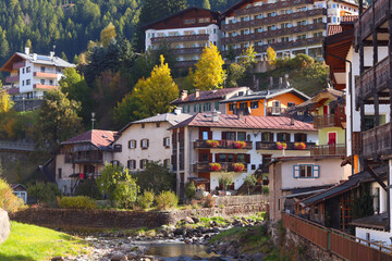 Picturesque autumn view of Moena, Italy - colorful alpine houses with flowered balconies along a mountain stream, surrounded by golden trees and sunlight in the Dolomites.