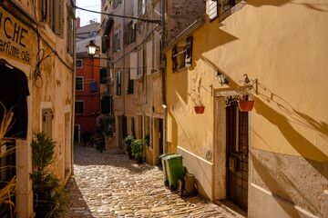Rovinj, Croatia - August 15, 2025: Golden sunlight fills a narrow cobblestone alley lined with pastel buildings, hanging plants, and shuttered windows, evoking a calm Mediterranean charm.