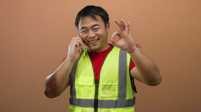 Man wearing yellow vest smiling while talking on smartphone against brown background showing ok gesture.