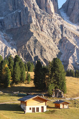 Cozy wooden chalets surrounded by pine trees at sunrise in the Alpe di Funes valley, with majestic Dolomite peaks rising in the background. Peaceful mountain landscape in autumn.