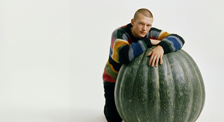 Young man with a striped sweater leaning on a large pumpkin in a minimalist setting