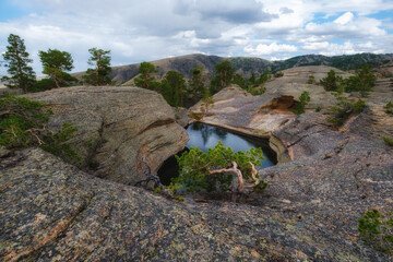 A mountainous landscape with a picturesque lake surrounded by rocky shores and sparse pine trees, located at the top of the Kent Mountains in Karkaraly Nature Park, this place is known as Forty Lakes.