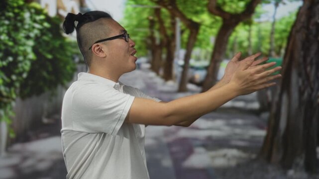 Man clapping hands on tree lined street, wearing white polo and glasses, ponytail undercut hairstyle and visible sleeves, looking to the right; joy welcome.