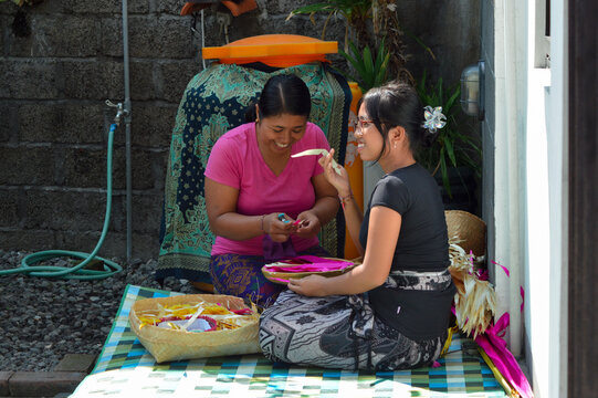 A mother and daughter sit on a mat outdoors, laughing and interacting happily while making Balinese offering crafts; the mother is smiling and the daughter is engaging sideways. - Powered by Adobe