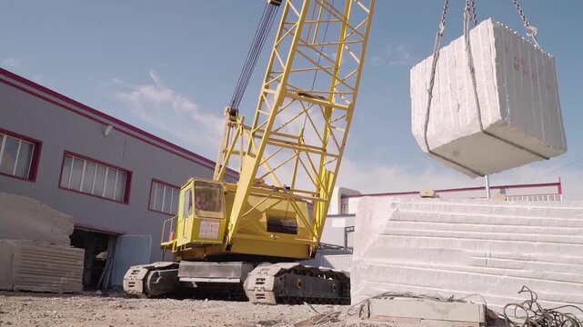 A yellow crawler crane lowers a granite block in the factory. Mining. Factory Building In the background. The tractor work in production. Stock. Granite slabs and blocks. Raw materials