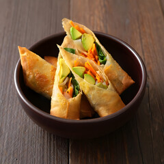 Crispy golden spring rolls filled with avocado carrots and greens served in a dark bowl on a wooden table