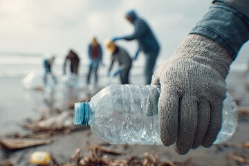 Gloved hand picks up a plastic bottle from a sandy beach with blurred cleaners in background