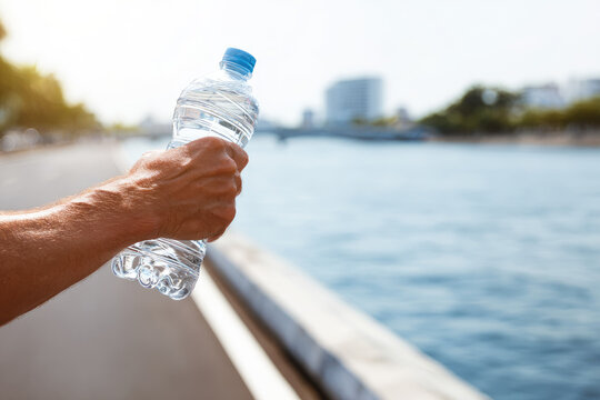 Close up of hand holding clear plastic water bottle during run along river, with city buildings and trees in background, evoking refreshing and active mood
