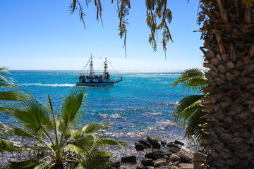 A picturesque view of a sailing ship navigating through vibrant turquoise waters, framed by lush palm trees under a clear blue sky on a sunny day.