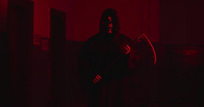 Grim figure of Death Reaper wearing black hooded cloak holding scythe and standing in dark hall dimly lit with sinister red light. Horror scene. Concept of death, evil, Halloween and scary stuff