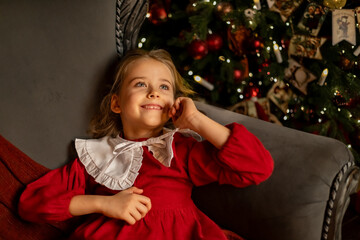 Little thoughtful girl lying on the sofa near Christmas tree in dark living room and looking up...