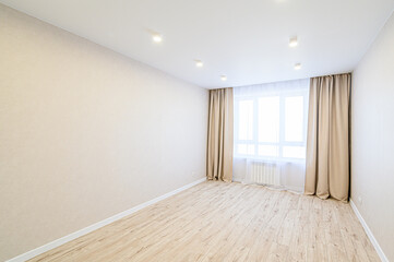 A well-lit, empty room with beige walls and light wooden floors. A bright window is flanked by beige curtains. Spotlights illuminate the ceiling