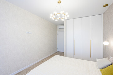 A bright, modern bedroom with white walls, hardwood floors, and a large wardrobe. The room is illuminated by a stylish chandelier and bedside lamp