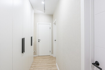 A modern hallway with white walls, light wood-look flooring, and white doors.  Features a white wardrobe with long black handles. Clean, minimalist design