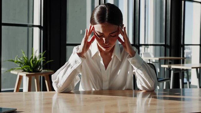 Woman holds head in pain. Professional employee sits at desk in office with window light. She shows headache and stress while rubbing her temples. Business woman looks tired and visibly overwhelmed. - Powered by Adobe