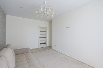 Bright, empty room with a beige sofa, white walls, and a modern chandelier. A paneled door stands out