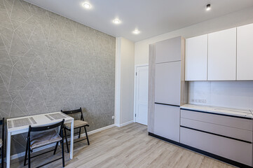 A modern kitchen area with a grey geometric wall, light wood flooring, white and grey cabinetry, a white table, and dark chairs. Minimalist style with recessed lighting