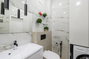 Modern bathroom featuring white marble-style tile, white sink, floating shelves, and a black toilet flush plate. Cylindrical pendant lights are also visible